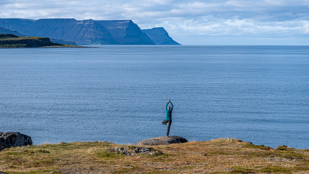 Figure of a woman in a yoga pose in front of the sea with mountains in the backgroun