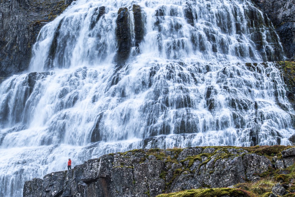 Small figure standing in front of a huge waterfall.
