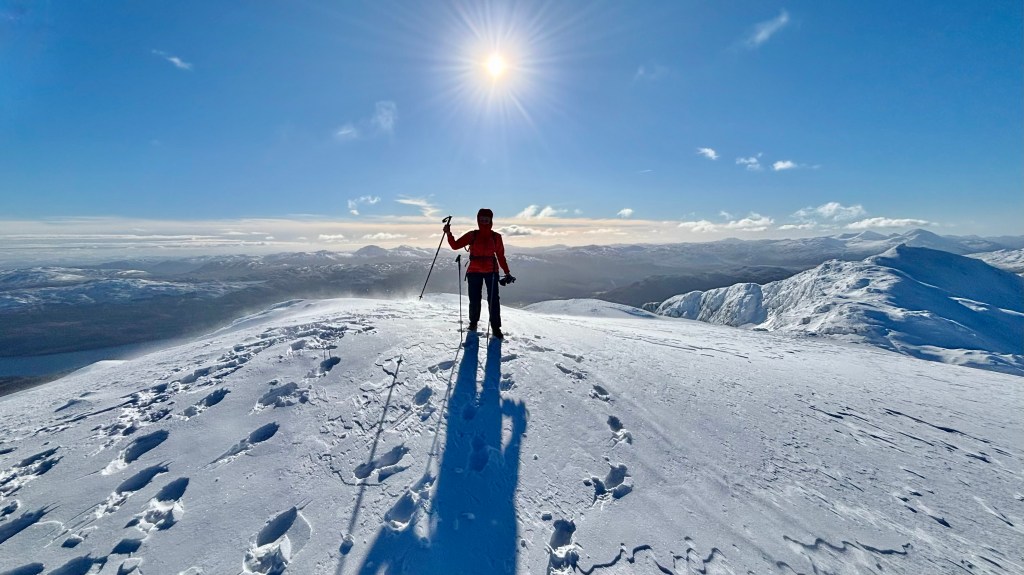 Female hiker stood on top of a snow covered mountain, with the sun behind her.