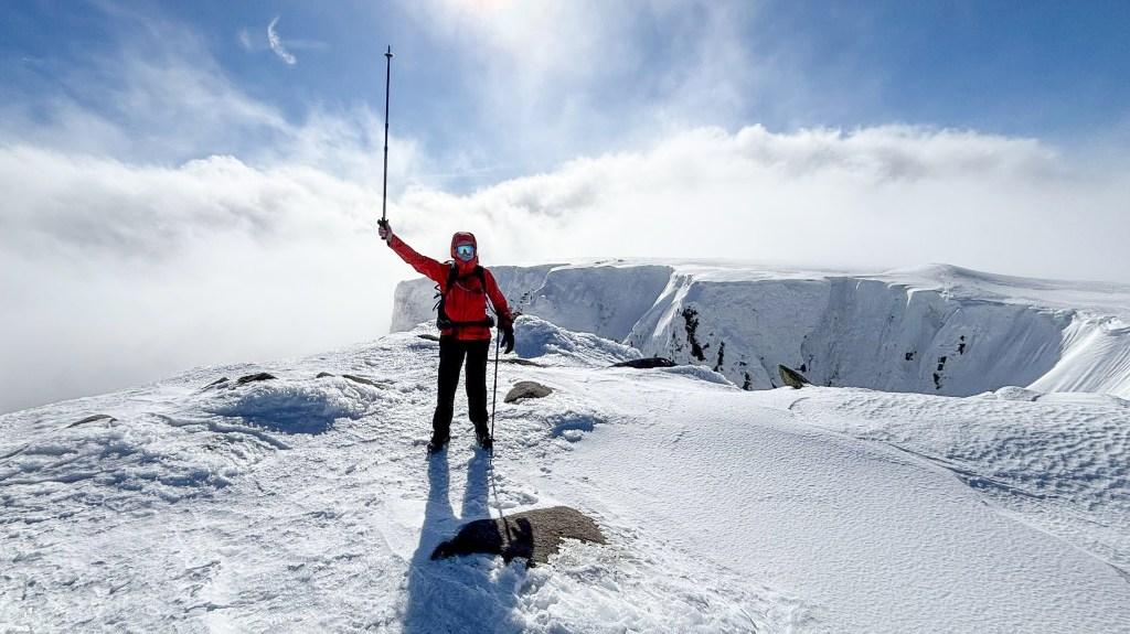 Female hiker stood on top of a snowy peak with her walking pole raised in her right hand.
