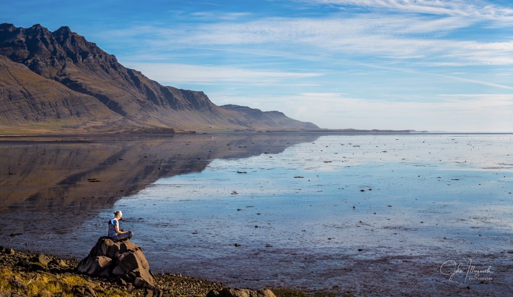 Figure sat meditating by a stretch of still water in Iceland.